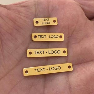 Puede incluir: Cuatro pequeñas etiquetas rectangulares de metal dorado con esquinas redondeadas y dos agujeros. Cada etiqueta tiene grabado el texto "TEXT - LOGO". Las etiquetas están dispuestas en una pila vertical, con la más pequeña arriba.