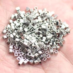 Puede incluir: Un montón grande de cuentas cuadradas de metal plateado con un pequeño agujero en cada cuenta. Las cuentas están esparcidas en una mano.