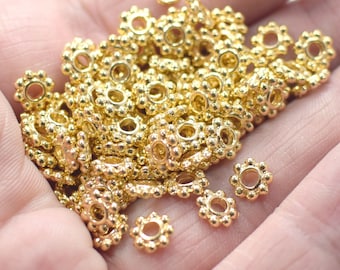 May include: A pile of gold-colored metal jump rings with a decorative flower design. The jump rings are small and have a round shape.