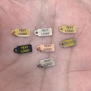 Könnte beinhalten: Kleine rechteckige Metallanhänger in Gold, Silber, Schwarz und Roségold. Jeder Anhänger hat ein kleines Loch und den Text "TEXT LOGO" in einer kontrastierenden Farbe. Die Anhänger sind wahrscheinlich für Schmuck- oder Bastelprojekte.