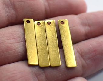 May include: Four gold-colored metal bar pendants with a hole at the top. The pendants are arranged in a row and are lying on a hand.