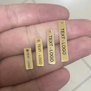 May include: Four small, rectangular gold-colored metal tags of varying sizes. Each tag has a small hole at the top and the text "TEXT-LOGO" printed in black. The tags are arranged in a row, with the smallest tag on the left and the largest on the right.