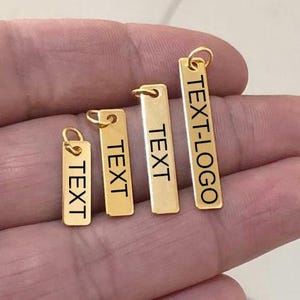Op de afbeelding: Vier goudkleurige rechthoekige hangers van verschillende formaten, elk met een kleine lus aan de bovenkant. De hangers zijn gegraveerd met het woord "TEXT" of "TEXT-LOGO" in zwart. De hangers worden in een hand gehouden.
