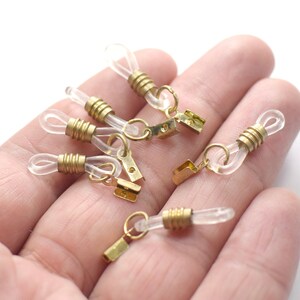 May include: A close-up of six clear plastic eyeglass chain holders with gold metal clasps. The holders are on a hand.