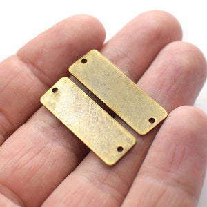 Puede incluir: Dos etiquetas rectangulares de metal de color dorado antiguo con dos agujeros cada una. Las etiquetas son ligeramente texturizadas y tienen un aspecto desgastado.