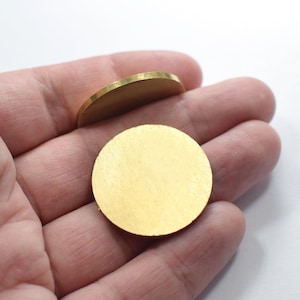 Puede incluir: Dos discos metálicos redondos de color dorado, uno encima del otro, en una mano.