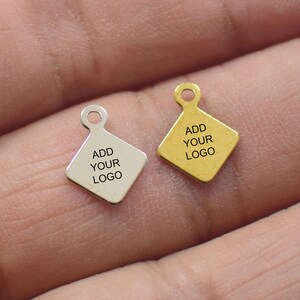 May include: Two diamond-shaped metal tags with a hole at the top. The tag on the left is silver and the tag on the right is gold. Both tags have the text "ADD YOUR LOGO" engraved on them.