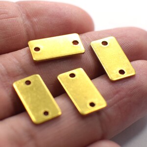 Pode incluir: Quatro etiquetas retangulares de metal de cor dourada com dois furos cada. As etiquetas estão em uma mão.