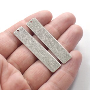 Könnte beinhalten: Zwei silberfarbene Metall-Bar-Anhänger mit strukturierter Oberfläche. Jeder Anhänger hat oben ein Loch zum Anbringen einer Kette oder Schnur.