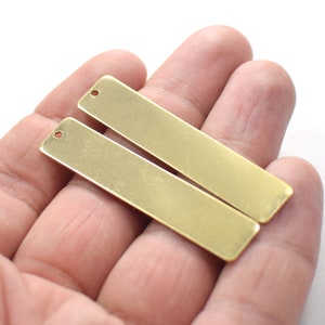 Könnte beinhalten: Zwei rechteckige Anhänger aus goldfarbenem Metall mit einem Loch oben. Die Anhänger liegen auf einer Hand.