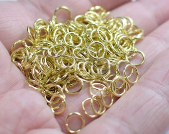 Puede incluir: Un montón grande de anillos de salto dorados, un tipo de hallazgo de joyería, en una mano. Los anillos de salto son pequeños y redondos, y son perfectos para conectar diferentes piezas de joyería.
