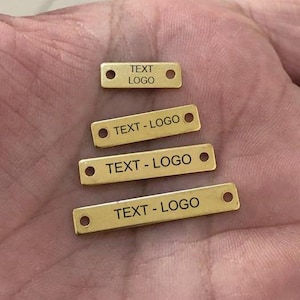 Puede incluir: Cuatro pequeñas etiquetas rectangulares de metal dorado con esquinas redondeadas y dos agujeros. Cada etiqueta tiene el texto "TEXT - LOGO" impreso en negro. La etiqueta superior dice "TEXT LOGO".