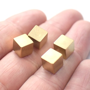 Puede incluir: Cuatro pequeños objetos metálicos de forma cúbica de color dorado. Los cubos están dispuestos en fila sobre la mano de una persona.
