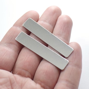 May include: Two silver metal rectangular bars, approximately 2 inches long, are held in a hand.