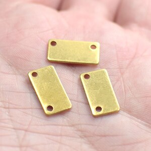 May include: Three gold-colored metal rectangular tags with two holes each. The tags are on a light pink surface.