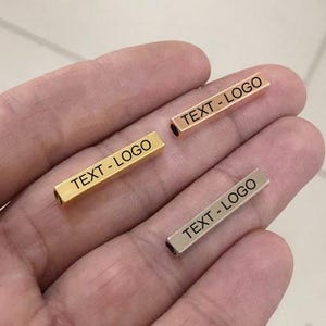 Puede incluir: Tres barras metálicas rectangulares en tonos oro, oro rosa y plata, cada una grabada con el texto "TEXT - LOGO". Las barras se sostienen en una mano, mostrando su acabado pulido y los detalles grabados.