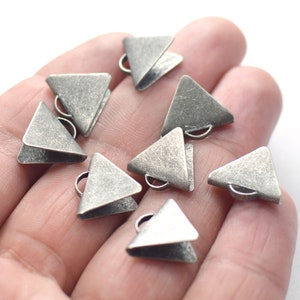 Puede incluir: Un grupo de ocho abalorios de metal de forma triangular plateados con un bucle en la parte superior. Los abalorios tienen un acabado texturizado y envejecido.