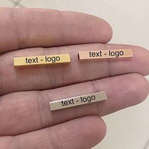May include: Three small rectangular bars in gold, rose gold, and silver, each with the text "text - logo" in black. The bars are held in a person's hand, showcasing their metallic finish and engraved text.