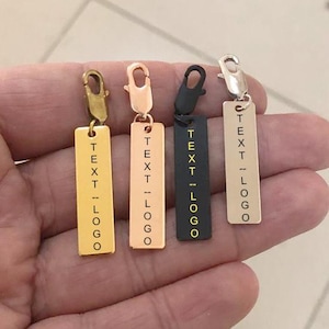 Puede incluir: Cuatro etiquetas de encanto de metal con cierres de mosquetón. Las etiquetas son doradas, oro rosa, negras y plateadas. Cada etiqueta tiene el texto "TEXT -- LOGO" en orientación vertical.