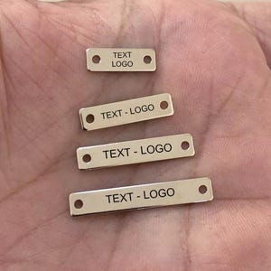 Puede incluir: Cuatro pequeñas etiquetas metálicas rectangulares con esquinas redondeadas. Cada etiqueta tiene un agujero en cada extremo. El texto "TEXT LOGO" está grabado en la etiqueta superior y "TEXT - LOGO" en las otras tres.
