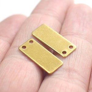 May include: Two gold-colored metal rectangular tags with two holes each. The tags are flat and have a smooth surface.