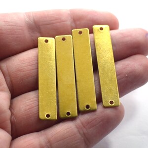 May include: Four gold-colored metal bar pendants with two holes each. The bars are smooth and have a slightly brushed finish.