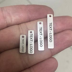Puede incluir: Cuatro pequeñas etiquetas rectangulares de metal de diferentes tamaños, cada una con un pequeño agujero en la parte superior. Las etiquetas son plateadas y presentan el texto "TEXT-LOGO" en negro. Las etiquetas se sostienen en una mano.