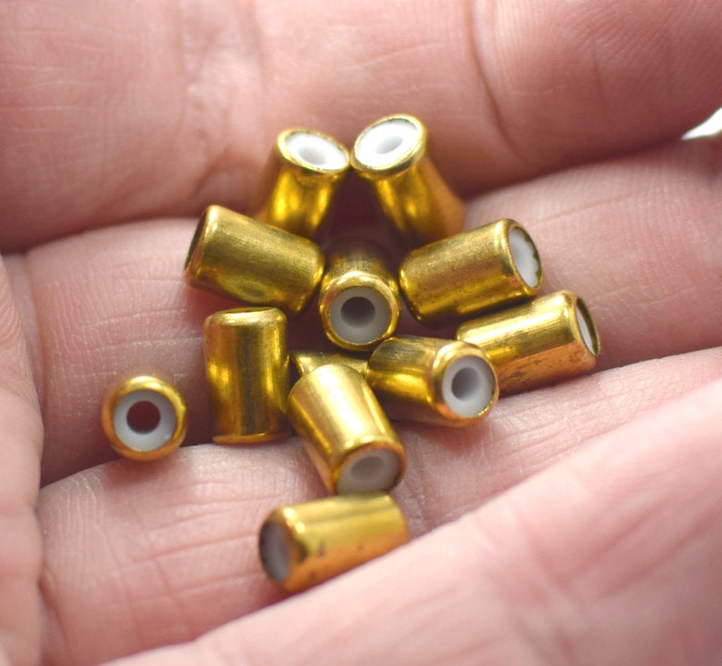 May include: A close-up of a pile of gold-coloured metal beads with white plastic inserts. The beads are cylindrical in shape and have a smooth, shiny finish.