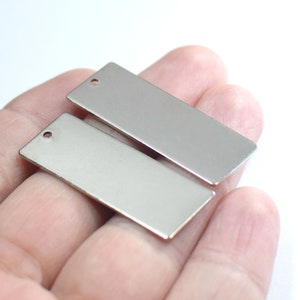 Pode incluir: Duas etiquetas retangulares de metal prateado com um furo no topo. As etiquetas estão sobre uma mão.