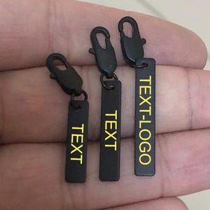 Op de afbeelding: Drie zwarte metalen ritstrekkers met een rechthoekig label. De labels hebben het woord "TEXT" of "TEXT-LOGO" in het geel gedrukt. De ritstrekkers zijn bevestigd aan een metalen sluiting.