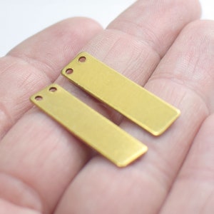 Pode incluir: Duas etiquetas retangulares de metal de cor dourada com dois furos cada. As etiquetas estão sobre uma mão.