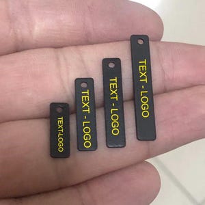 Peut inclure: Quatre étiquettes rectangulaires noires de différentes tailles, chacune avec un petit trou en haut. Le texte "TEXT-LOGO" est imprimé en jaune sur chaque étiquette. Les étiquettes sont présentées sur un fond neutre.