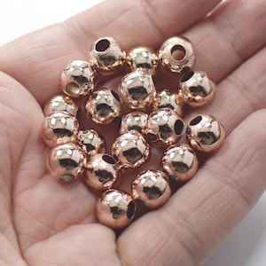 May include: A collection of rose gold-colored metal beads. Each bead is round with a textured surface and a hole for stringing. The beads have a shiny, reflective finish, and are held in a hand.