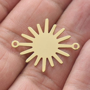 May include: A gold-colored sunburst charm with a central disc and radiating rays. The charm has two small loops extending from the sides. This jewelry component is approximately 1 inch in diameter.