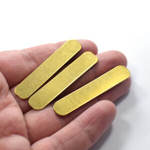 Pode incluir: Três retângulos de metal de cor dourada, possivelmente para a fabricação de joias, são mantidos em uma mão.