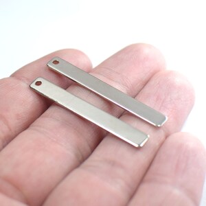 Puede incluir: Dos colgantes de barra de metal plateado con un agujero en la parte superior. Los colgantes están sobre una mano.