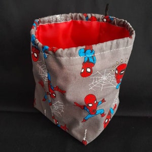 K&ouml;nnte beinhalten: Ein grauer Stoffw&uuml;rfelbeutel mit roter Innenausstattung. Der Beutel ist mit einem sich wiederholenden Muster von Spiderman-Charakteren verziert.