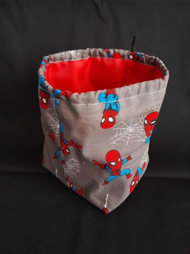 K&ouml;nnte beinhalten: Ein grauer Stoffw&uuml;rfelbeutel mit roter Innenausstattung. Der Beutel ist mit einem sich wiederholenden Muster von Spiderman-Charakteren verziert.