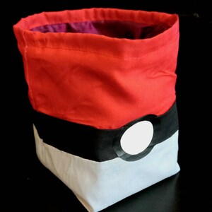 K&ouml;nnte beinhalten: Roter, schwarzer und wei&szlig;er Stoff-W&uuml;rfelbeutel mit einem Pokeball-Design. Die Tasche hat einen Kordelverschluss.