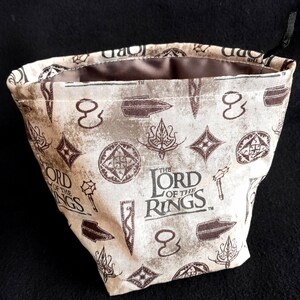 K&ouml;nnte beinhalten: Ein beigefarbener W&uuml;rfelbeutel aus Stoff mit einem braunen Kordelzug. Der Beutel ist mit einem sich wiederholenden Muster des "Der Herr der Ringe"-Logos, Schwertern und anderen Symbolen bedruckt.