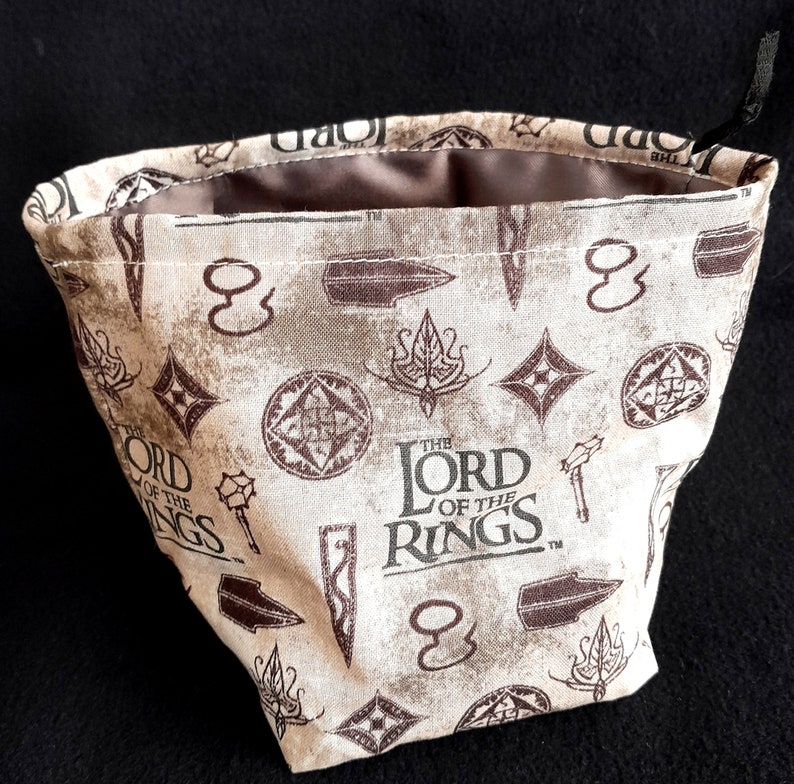 K&ouml;nnte beinhalten: Ein beigefarbener W&uuml;rfelbeutel aus Stoff mit einem braunen Kordelzug. Der Beutel ist mit einem sich wiederholenden Muster des "Der Herr der Ringe"-Logos, Schwertern und anderen Symbolen bedruckt.