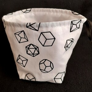K&ouml;nnte beinhalten: Ein wei&szlig;er Stoff-W&uuml;rfelbeutel mit Kordelzug. Der Beutel ist mit schwarzen geometrischen Formen bedruckt, die W&uuml;rfeln &auml;hneln.