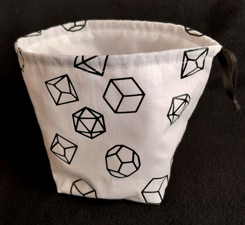 K&ouml;nnte beinhalten: Ein wei&szlig;er Stoff-W&uuml;rfelbeutel mit Kordelzug. Der Beutel ist mit schwarzen geometrischen Formen bedruckt, die W&uuml;rfeln &auml;hneln.