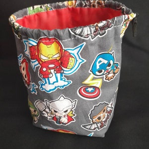 K&ouml;nnte beinhalten: Ein grauer Stoff-W&uuml;rfelbeutel mit Kordelzug. Der Beutel ist mit einem sich wiederholenden Muster von Comic-Superhelden verziert, darunter Iron Man, Captain America, Thor und andere. Der Beutel hat ein rotes Futter.