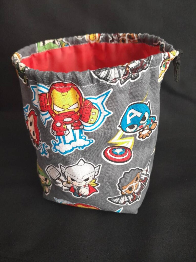 K&ouml;nnte beinhalten: Ein grauer Stoff-W&uuml;rfelbeutel mit Kordelzug. Der Beutel ist mit einem sich wiederholenden Muster von Comic-Superhelden verziert, darunter Iron Man, Captain America, Thor und andere. Der Beutel hat ein rotes Futter.