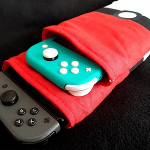 May include: A red and black fabric pouch with a white circle design. A teal Nintendo Switch Joy-Con controller is inside the pouch.