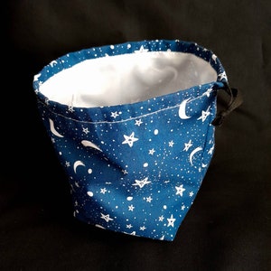 K&ouml;nnte beinhalten: Eine blaue Stofftasche mit Kordelzug und einem wei&szlig;en Muster aus Mondsicheln und Sternen. Die Tasche ist leer und der Kordelzug ist festgezogen.