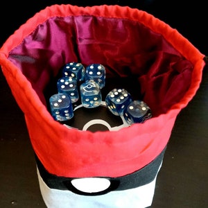 K&ouml;nnte beinhalten: Roter und schwarzer Stoff-W&uuml;rfelbeutel mit einem wei&szlig;en Pokeball-Design. Der Beutel ist mit blauen W&uuml;rfeln gef&uuml;llt.