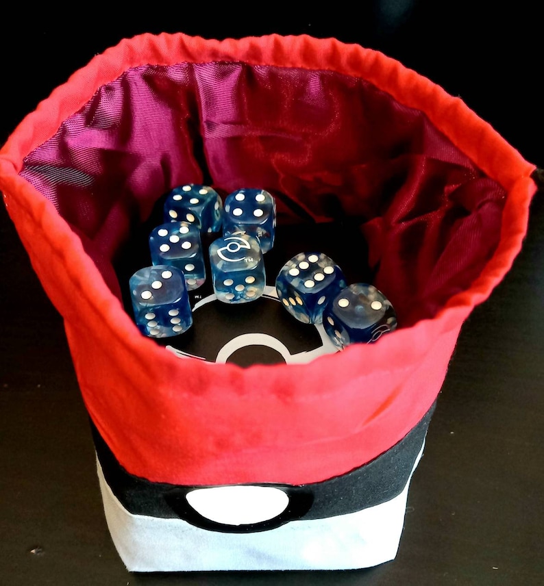 K&ouml;nnte beinhalten: Roter und schwarzer Stoff-W&uuml;rfelbeutel mit einem wei&szlig;en Pokeball-Design. Der Beutel ist mit blauen W&uuml;rfeln gef&uuml;llt.