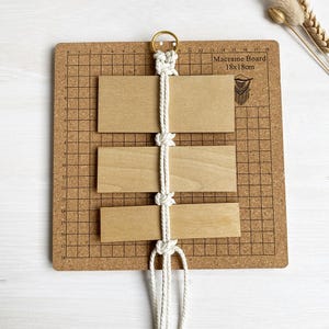 Peut inclure: Un cordon de macramé blanc est enfilé à travers trois planches de bois sur un panneau en liège avec un motif de grille. Les planches mesurent 18 cm sur 18 cm. Le cordon de macramé est noué et pend sous les planches.